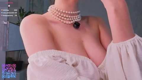 Video of a light-skinned woman with a slender physique, wearing a beige off-shoulder top and a pearl necklace. Background shows indoor setting with a potted plant.