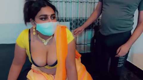 Video of a South Asian woman in a yellow blouse, exposing cleavage, with a blue face mask, sitting on a bed in a room with a man in the background.
