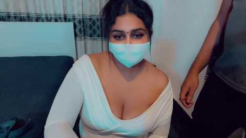 Video of a South Asian woman with a medium complexion, wearing a white off-shoulder top, blue surgical mask, and glasses, seated on a dark couch, with a man in a gray shirt standing behind her.