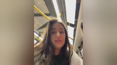 Video of a young woman with long brown hair, wearing a black top, seated in a public bus. The background shows yellow handrails and a reflective window.