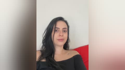 Video of a young woman with long black hair, wearing a black off-shoulder top, sitting against a white wall with a red cushion behind her.