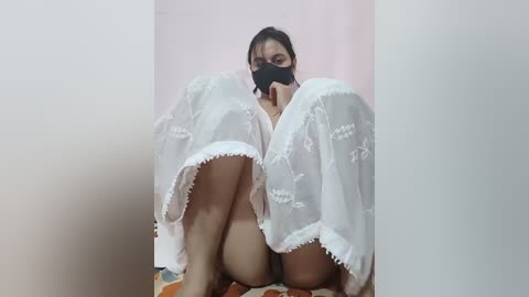 Video of a woman with short black hair, wearing a black face mask, a sheer white nightgown, and white lace panties, crouched in a corner, legs bent, with a pastel-colored wall behind her.