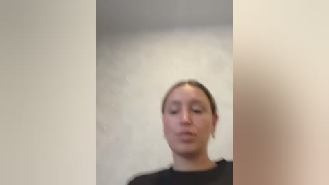 A video shows a woman with light skin and brown hair tied back, wearing a black top, standing in a plain, light-colored room. The image is slightly blurred.