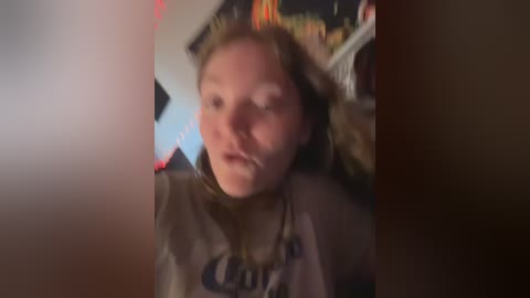 Media: A video of a young woman with light skin and long brown hair, wearing a grey shirt, with a blurred background featuring red and blue lights.