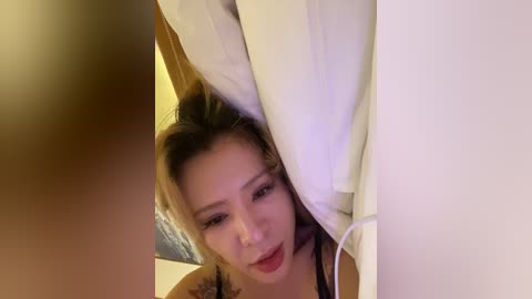 Video of an Asian woman with blonde hair, partially hidden by white pillows, wearing a black top, with a tattoo visible on her left arm.