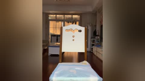 Video of a modern, sunlit living room with hardwood floors, featuring a white, upholstered armchair with three round buttons on its backrest, and a baby's crib with a white, patterned sheet in the foreground.