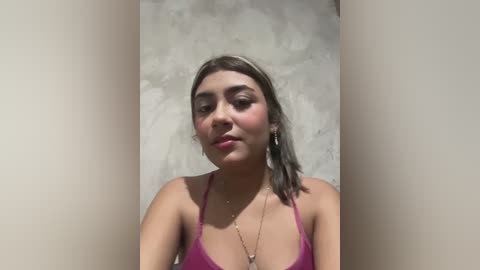 Video of a young woman with light brown skin, wearing a pink tank top, standing against a textured, off-white wall. She has straight, shoulder-length hair, minimal makeup, and a neutral expression.