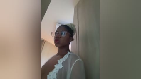 Video of a black woman with short, blonde hair in glasses, wearing a sheer, white, frilled robe, standing in a dimly lit room with beige walls and a slanted ceiling.