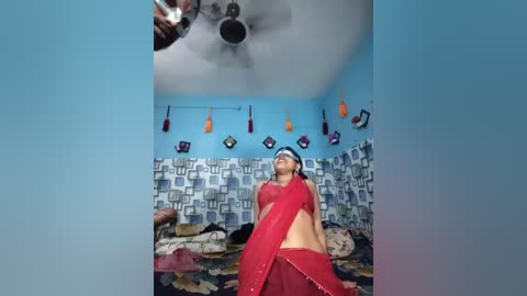 A video of a woman in a red sari, blindfolded, standing in a dimly lit room with blue and gray patterned wallpaper and a fan.