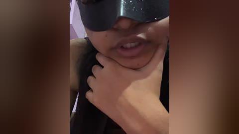 A close-up video of a woman with medium brown skin, wearing a black mask, and holding a man's penis. She has a neutral facial expression and is partially obscured by the mask.