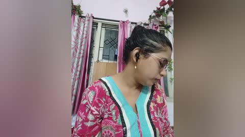 Video of a South Asian woman in a floral pink and blue hijab, standing in a room with pink curtains and a window, looking down.