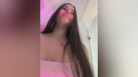 Video of a young woman with long brown hair, wearing a white strapless top, smiling, in a dimly lit room with pink and purple lighting.