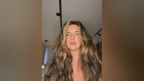 Video of a fair-skinned woman with long, wavy, blonde hair, wearing red lipstick, indoors in a modern, minimally decorated room with a white ceiling and black track lighting.