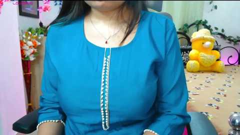 Video of a woman with medium brown skin wearing a blue long-sleeve blouse with white embroidery, sitting indoors with a yellow teddy bear, a table, and floral decor.