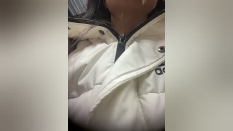 A close-up video of a woman in a white puffer jacket with a black zipper, showing her partially open jacket and a visible drop of liquid on her chin. The background is blurred, focusing on her upper body.