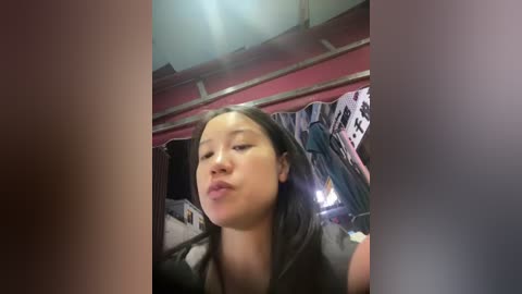 Video of a young Asian woman with long black hair, pouting in a cluttered, dimly-lit room with a red wall, clothes hanging on racks, and a ceiling light.