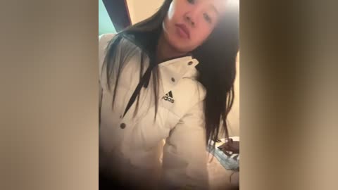 A video of an Asian woman with long black hair, wearing a white Adidas jacket, captured in a dimly lit indoor setting.