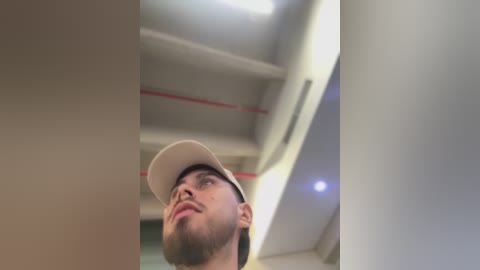 Video of a young man with a light complexion and trimmed beard, wearing a white cap, looking up thoughtfully under fluorescent lighting in a modern, well-lit room.
