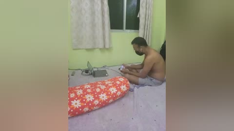 Video of a shirtless, dark-skinned man with a muscular build sitting on a bed, holding a remote, in a modestly furnished room with pale yellow walls and a white window.