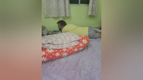 Video of a man lying on a bed with a red floral blanket, wearing a yellow shirt and gray pants, in a room with green walls and a white curtain.