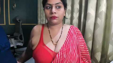 Video of a dark-skinned woman with short black hair, wearing a red and white striped sari, a gold necklace, and red lipstick, in a dimly lit room with green walls and patterned curtains.