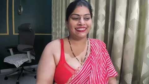 Video of a South Asian woman with dark skin, black hair in a bun, smiling, wearing a red and white striped saree, and a gold necklace. Background features a dark green wall, office chair, and beige curtains.