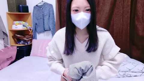 Video of a young Asian woman with long black hair and a white mask, wearing a cream sweater, sitting on a bed in a room with clothes on shelves, brown curtains, and a pink shopping bag.