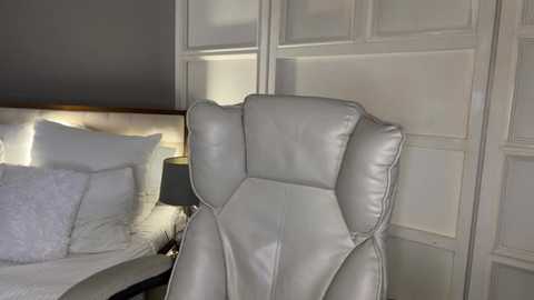 Video of a modern bedroom featuring a plush, white leather recliner chair, a neatly made bed with white bedding, and a white-paneled wall with frosted glass doors.