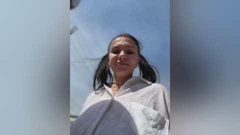 A video of a young woman with light skin and black pigtails, wearing a white, loose-fitting shirt, captured from a low angle. The background features a clear blue sky.