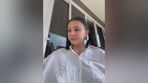 Video of a young woman with fair skin, black hair in pigtails, wearing a white shirt, pearl necklace, and earrings, standing in a modern room with white walls and glass doors.