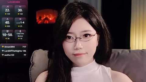 Video of a young East Asian woman with long black hair, wearing glasses, a white sleeveless top, and sitting on a couch in a dimly lit room.