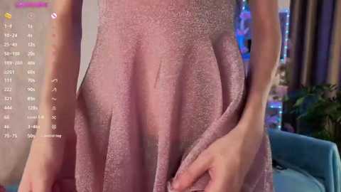 A close-up video of a light-skinned person wearing a pink, textured dress, standing in a room with a blue couch, a plant, and a TV screen displaying a digital clock and temperature.