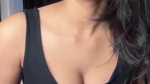 Video of a woman's chest in a black tank top, showcasing a moderate cleavage, with wavy dark hair partially covering her neck. The background is blurry, featuring dark, possibly black curtains.