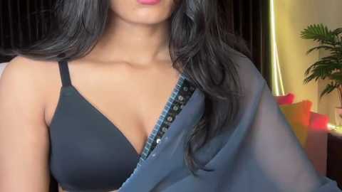 Video of a South Asian woman with medium-brown skin, long black hair, wearing a black bra and a blue see-through sari, with a plant and colorful pillows in the background.