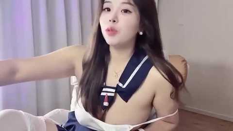 Video of an Asian woman with long dark hair, wearing a white sailor dress with a navy blue collar, sitting on a white chair, making a kissing gesture.