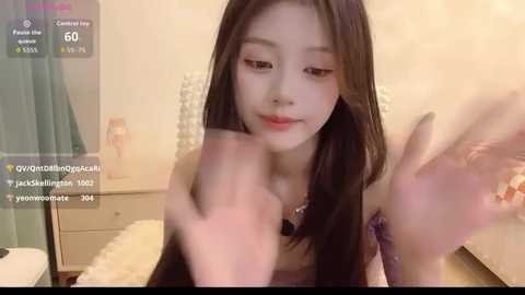 A video of an Asian woman with long, straight brown hair and fair skin, wearing a purple top, sitting on a white chair. She's holding a phone, and the background shows a bedroom with a bed and beige walls.