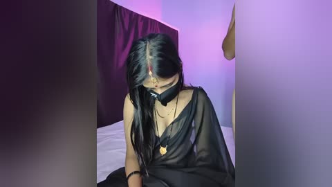 Video of a young woman with long black hair, wearing a sheer black dress, a gold necklace, and a black face mask, seated in a dimly lit room with purple and blue lighting.