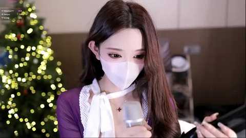 Video of an Asian woman with long brown hair, wearing a white face mask, purple shirt, and white lace collar, reading a book, standing in a softly lit, festive room with a decorated Christmas tree in the background.