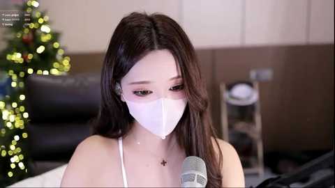 Video of an East Asian woman with long, straight brown hair, wearing a white face mask, sitting at a desk with a Christmas tree and festive lights in the background.