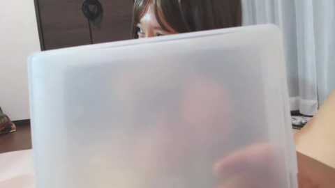 A video of a woman with long brown hair partially obscured by a clear plastic bag, revealing her face and shoulder, in a modern room with light-colored walls, a window with white curtains, and a wooden dresser.
