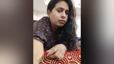 Video of a South Asian woman with long black hair, wearing a dark patterned top, lying on a red and white patterned blanket.