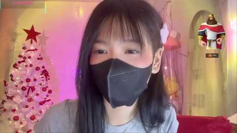 A video of a young Asian woman with straight black hair, wearing a black face mask, gray shirt, and a white hair clip, in a festive room with Christmas decorations, a tree, and Santa Claus figurine.