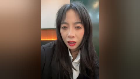 Video of an Asian woman with long, straight black hair, wearing a black blazer over a white shirt, smiling brightly. Background features a blurred, orange-tiled wall.