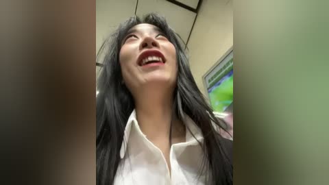 Video of an Asian woman with long black hair, wearing a white blouse, laughing with her mouth wide open, taken from a low angle. Background shows a TV and ceiling tiles.