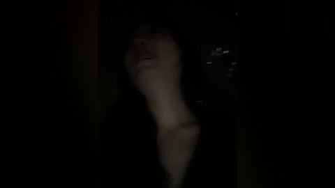 Video of a dimly lit, shadowy figure with a blurred face, possibly female, standing against a dark, possibly urban background with faintly visible city lights.
