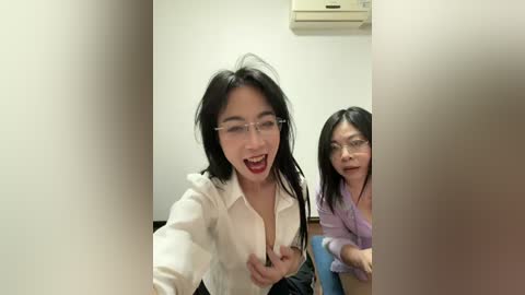 Video of two Asian women in an office. One with long black hair, glasses, and a white blouse, playfully sticking her tongue out. Another with short hair, glasses, and a purple shirt, looking amused.