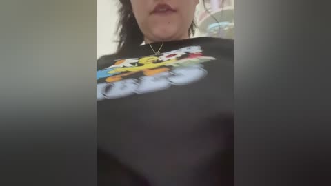 Video of a woman's upper body wearing a black graphic tee with a colorful, cartoonish design featuring characters in action poses. The background is blurred, with hints of a light-colored wall and a colorful object.