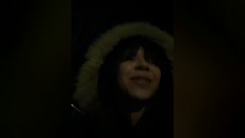 A dimly lit video of a woman with long black hair and a fur-lined hood, smiling slightly, set against a dark, blurry background.