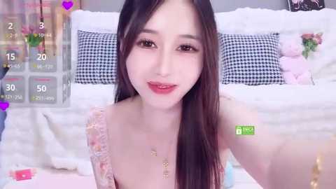 Video of an East Asian woman with long black hair, wearing a pink lace dress, smiling in a bedroom with white bedding and black-and-white pillows.