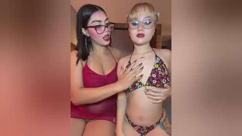 Video of two women: one with long black hair, red top, and glasses, and the other with short blonde hair, floral bikini, and glasses, both smiling.